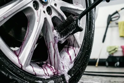 Cerca de rueda de BMW con llantas plateadas contaminadas por hierro oxidado púrpura durante descontaminación: brocha negra aplicando limpiador con espuma púrpura abundante, hidrolimpiadora de fondo en taller de lavado autos.