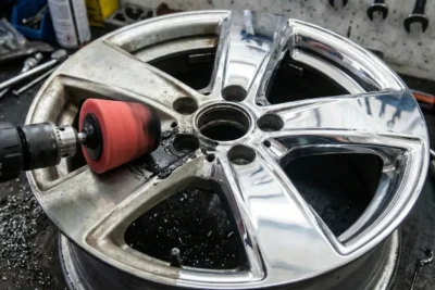 Mano con guantes usando taladro con cepillo rojo de goma puliendo llanta de aleación plateada oxidada en taller mecánico, eliminando suciedad y restaurando brillo metálico